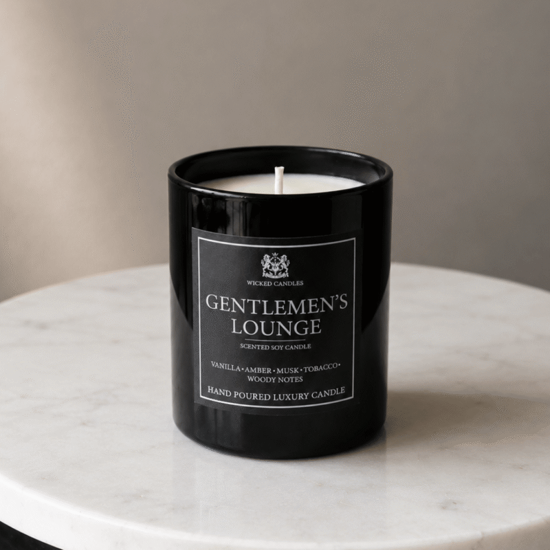Luxury Gentleman’s Lounge αρωματικό κερί σόγιας της Wicked Candles με νότες vanilla, amber, musk, tobacco και woody notes σε glossy μαύρο γυάλινο βάζο με φλόγα πάνω σε σκούρο ξύλινο desk background και warm moody luxury φωτισμό.